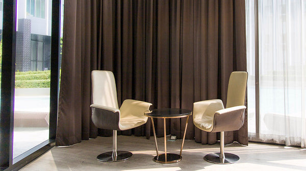 A sophisticated office lounge or hotel reception area featuring custom full-length brown blackout curtains, modern cream-colored swivel chairs, and a minimalist side table.